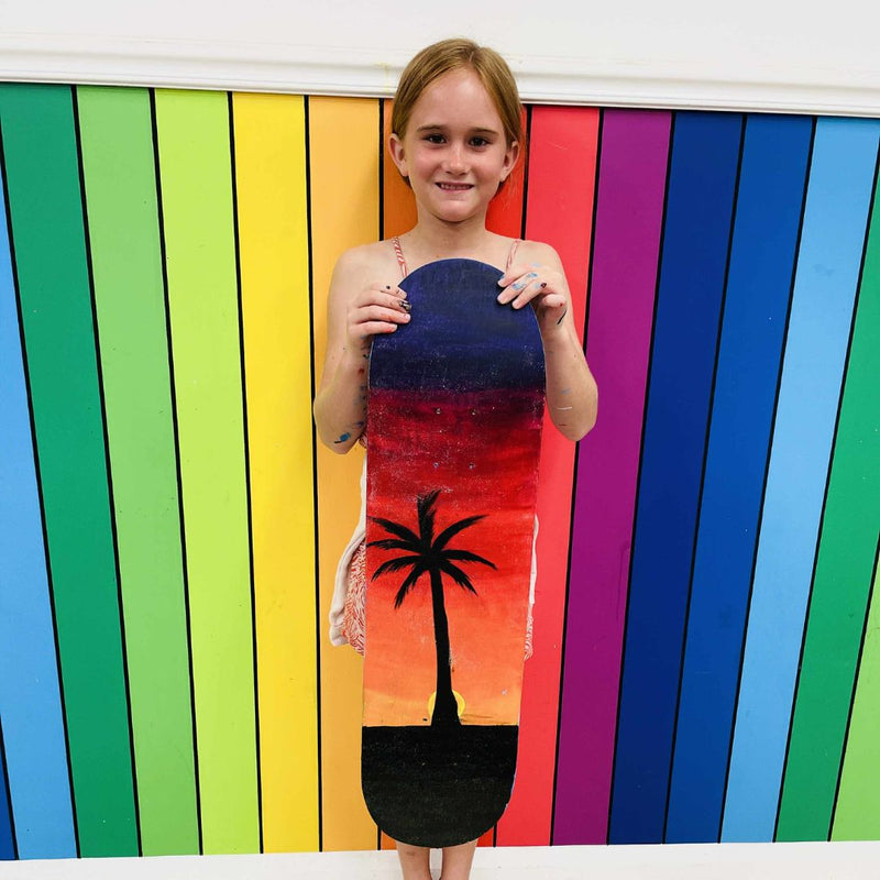 Girl holding a skateboard deck with a palm tree design in DIY Invasion workshop room.