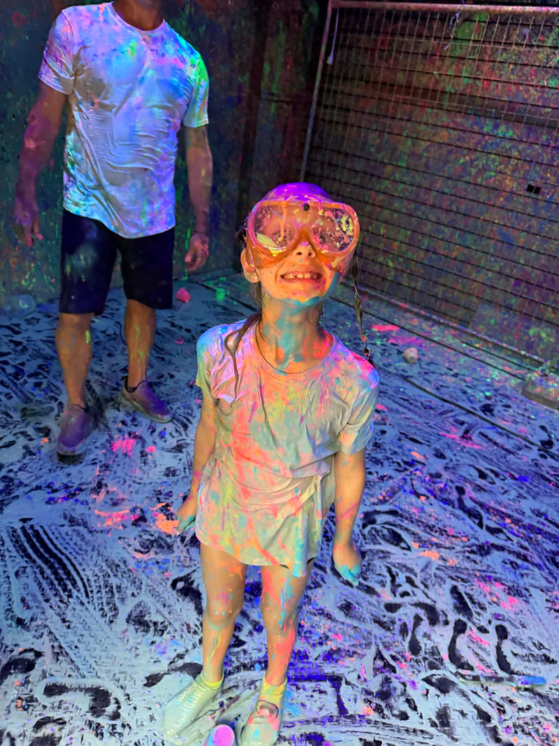 Child wearing goggles and standing on a floor with paint splashes, next to an adult.